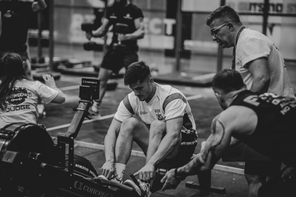Sportif au rameur pendant une compétition de fitness, encouragé de près par deux personnes, une juge à côté, photo en noir et blanc